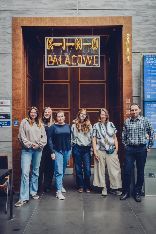 Sześcioro uczestników wizyty studyjnej pozuje przed drewnianymi drzwiami sali kinowej. Nad drzwiami neon z napisem „Kino Pałacowe”, obok świetlne oznaczenie „Sala 1”.