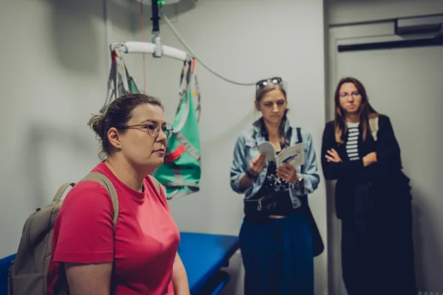 Pomieszczenie higieniczne z sufitowym podnośnikiem i zielonym nosidłem oraz niebieską leżanką. Trzy uczestniczki wizyty oglądają wyposażenie, jedna z nich czyta drukowany program spotkania.