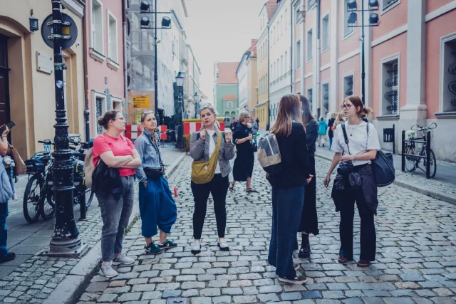 Grupa uczestniczek wizyty studyjnej stoi na nierównej, brukowanej uliczce wśród kolorowych kamienic. Kobieta pośrodku, z żółtą torbą na ramieniu, mówi do pozostałych.