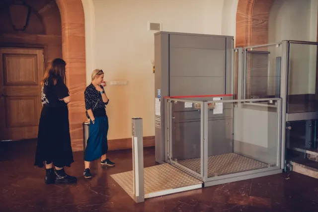 Dwie uczestniczki wizyty oglądają podnośnik pionowy dla osób poruszających się na wózku, zamontowany przy schodach w zabytkowym wnętrzu.