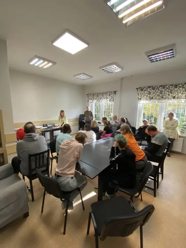 Kilkanaście osób siedzi przy dużym stole w jasnej sali, słuchając osoby prowadzącej stojącej przy stole na końcu sali. W pomieszczeniu są okna z zasłonami w roślinny wzór, a światło pada przez duże, prostokątne lampy na suficie.