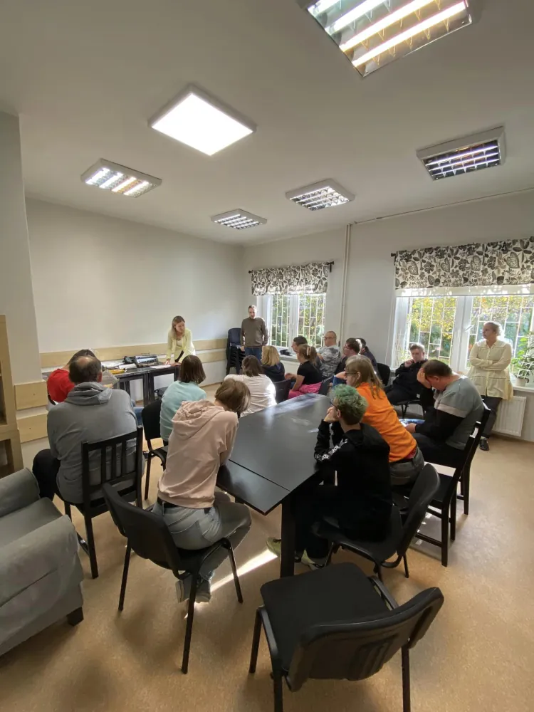 Kilkanaście osób siedzi przy dużym stole w jasnej sali, słuchając osoby prowadzącej stojącej przy stole na końcu sali. W pomieszczeniu są okna z zasłonami w roślinny wzór, a światło pada przez duże, prostokątne lampy na suficie.