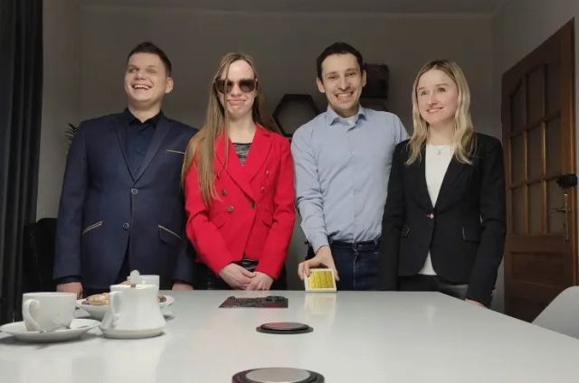 Na fotografii widać czwórkę ludzi stojących za białym stołem w jasnym pomieszczeniu. Od lewej strony: mężczyzna w ciemnoniebieskim garniturze z uśmiechem na twarzy, kobieta w czerwonej marynarce i czarnych spodniach z ciemnymi okularami przeciwsłonecznymi, mężczyzna w niebieskiej koszuli i ciemnych spodniach, oraz kobieta w czarnej marynarce i białej bluzce. Wszyscy są ubrani elegancko, a ich postawy są wyprostowane i uśmiechnięte, co może sugerować formalne lub półformalne spotkanie. Na stole widoczne są elementy takie jak filiżanki i dzbanek, co może wskazywać na przerwę na kawę lub herbatę.