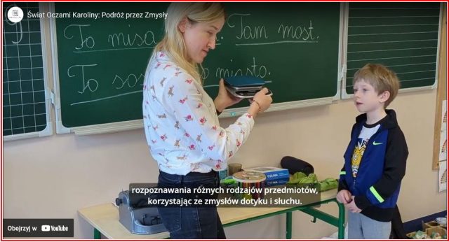 Na zdjęciu widzimy młodą kobietę i chłopca w klasie szkolnej. Kobieta, ubrana w białą koszulę w różowe jednorożce, stoi przy drewnianym biurku, na którym znajdują się różne przedmioty. Trzyma w rękach czarne urządzenie, które wygląda na maszynę do pisania dla osób niewidomych. Obok na biurku widać puszki i inne przedmioty, które mogą być używane do ćwiczeń sensorycznych. W tle widać zieloną tablicę, na której napisano kredą po polsku "To misa", "To słoik", "To mięta" oraz "Tam most". Chłopiec, stojący obok kobiety, ubrany jest w ciemną bluzę z kapturem i patrzy w stronę kobiety, być może oczekując na instrukcje lub uczestnicząc w zajęciach. Na dole zdjęcia znajduje się opis w języku polskim, który wspomina o rozpoznawaniu różnych rodzajów przedmiotów, korzystając ze zmysłów dotyku i słuchu.