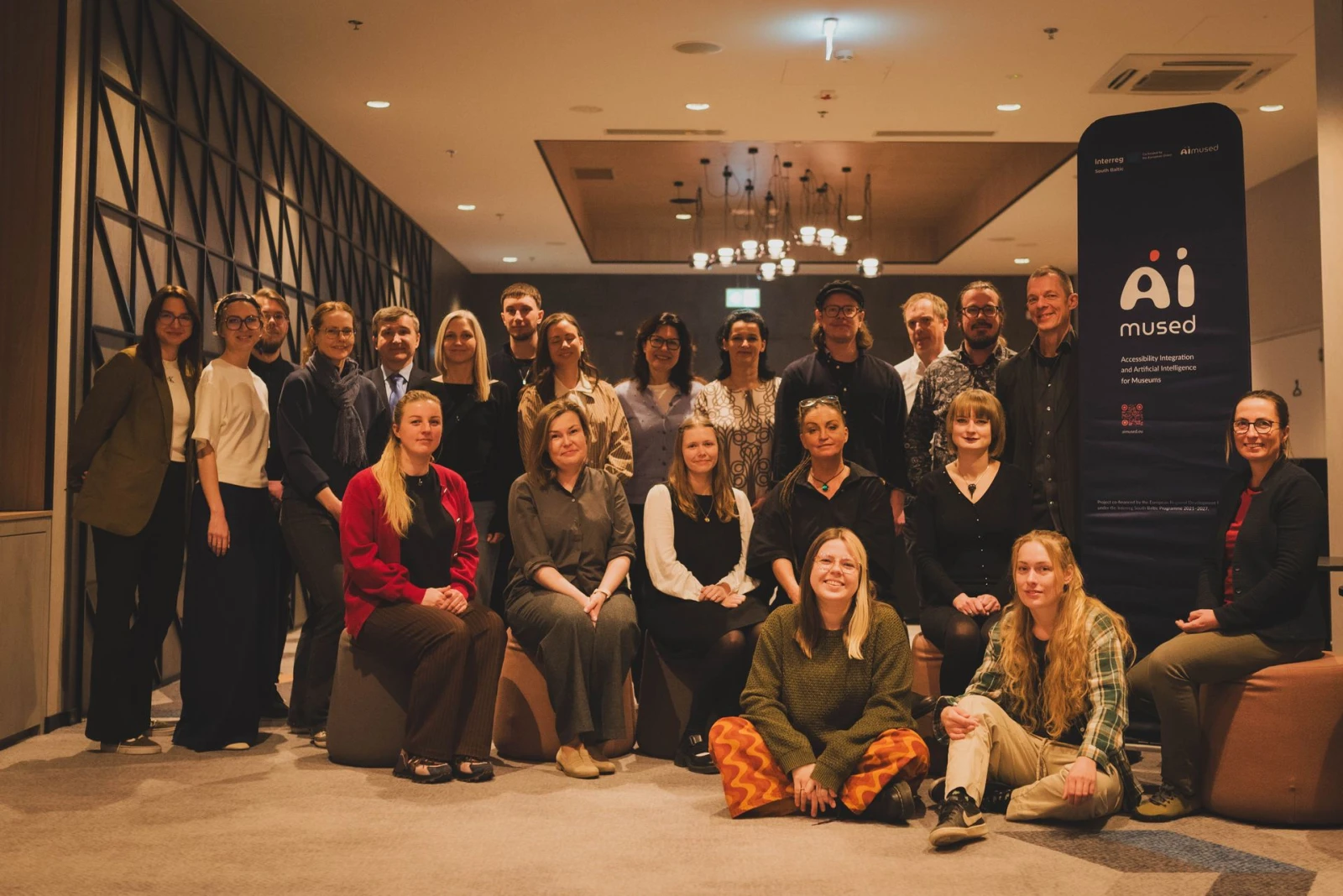 Zdjęcie grupowe uczestników spotkania projektowego we wnętrzu. Po prawej stronie roll-up z logo AIMused oraz Interreg Południowy Bałtyk i informacją o współfinansowaniu przez Unię Europejską.