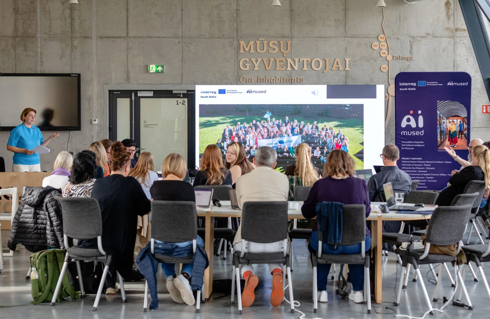 Sala konferencyjna podczas spotkania projektu. Prelegentka stoi przy ekranie ze zdjęciem grupowym i logotypami Interreg oraz AIMused. Na ścianie napis „Mūsų gyventojai – Our inhabitants”, po prawej roll-up z listą partnerów projektu. Uczestnicy siedzą przy stołach tyłem do obiektywu.