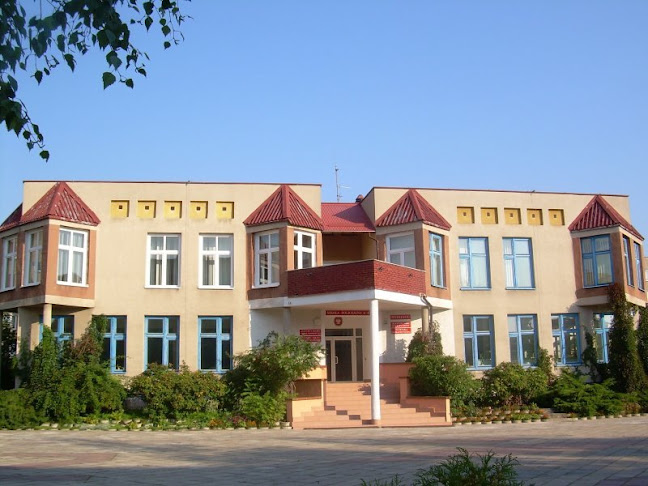 Elewacja dwukondygnacyjnego, żółto‑beżowego budynku urzędu gminy z centralnym wejściem przez schody pod balkonem, otoczonego krzewami i licznymi oknami.