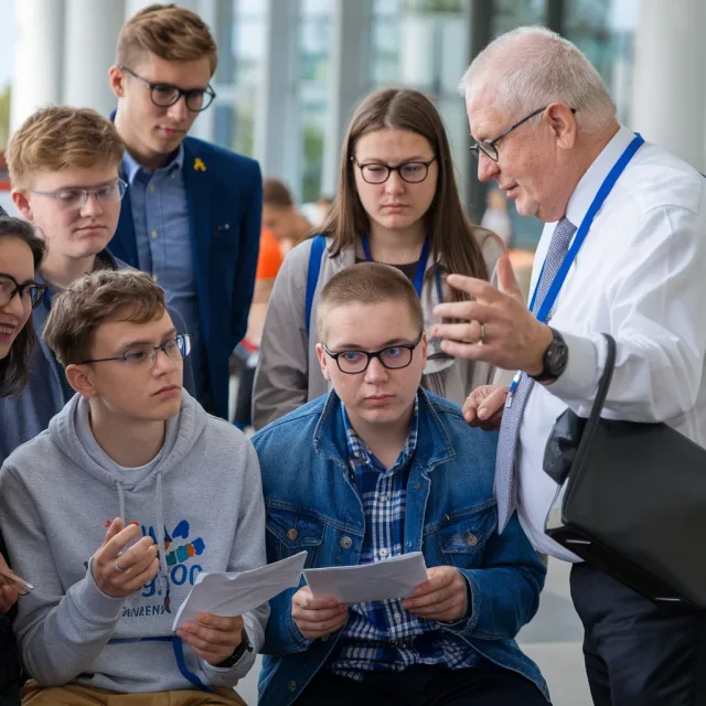 Na zdjęciu widać grupę młodych osób, zarówno chłopców, jak i dziewcząt, słuchających uważnie starszego mężczyzny w okularach, ubranego w białą koszulę i krawat. Mężczyzna gestykuluje, prawdopodobnie tłumacząc coś lub udzielając wskazówek. Dwóch młodych chłopaków siedzi z przodu, trzymając kartki papieru i patrząc z koncentracją na rozmówcę. W tle widoczne są przeszklone ściany budynku oraz inne osoby. Atmosfera zdjęcia sugeruje edukacyjne lub informacyjne spotkanie, być może warsztaty lub konsultacje.