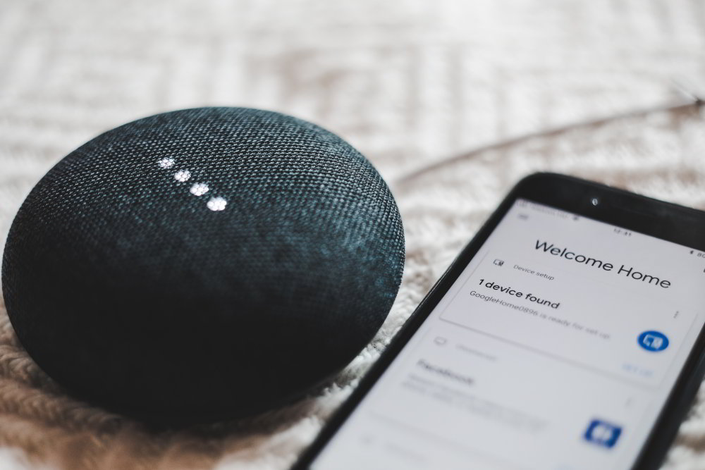 Zdjęcie przedstawia głośnik przenośny leżący obok telefonu komurkowego z włączoną aplikacją smarthome.