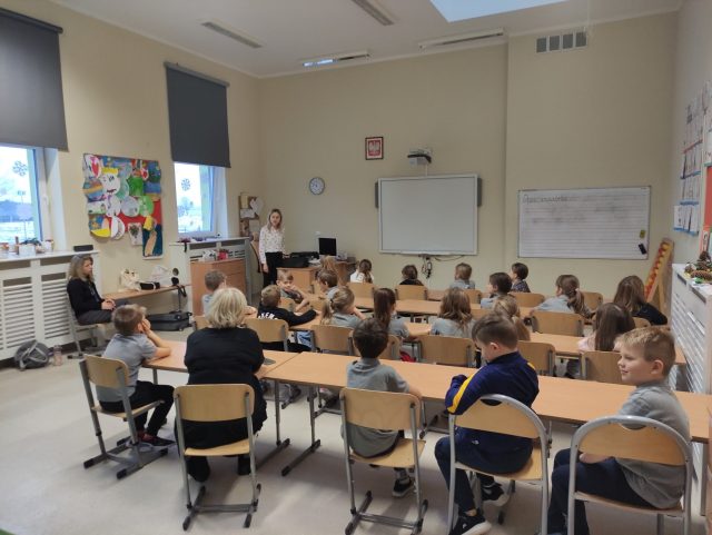 Na zdjęciu widać klasę szkolną podczas zajęć. Uczniowie w wieku szkolnym podstawowym siedzą na drewnianych krzesłach skierowanych w stronę białej tablicy, na której nie ma żadnych zapisków. Są rozmieszczeni w uformowanych rzędach, skupieni na osobie stojącej przed klasą. Po prawej stronie klasy znajduje się kącik z kolorowymi pracami plastycznymi uczniów i tablica z zapisem lekcji. Po lewej stronie widać okna z zasłonami oraz zegar na ścianie. Klasa jest dobrze oświetlona, a atmosfera wydaje się być spokojna i zorganizowana.