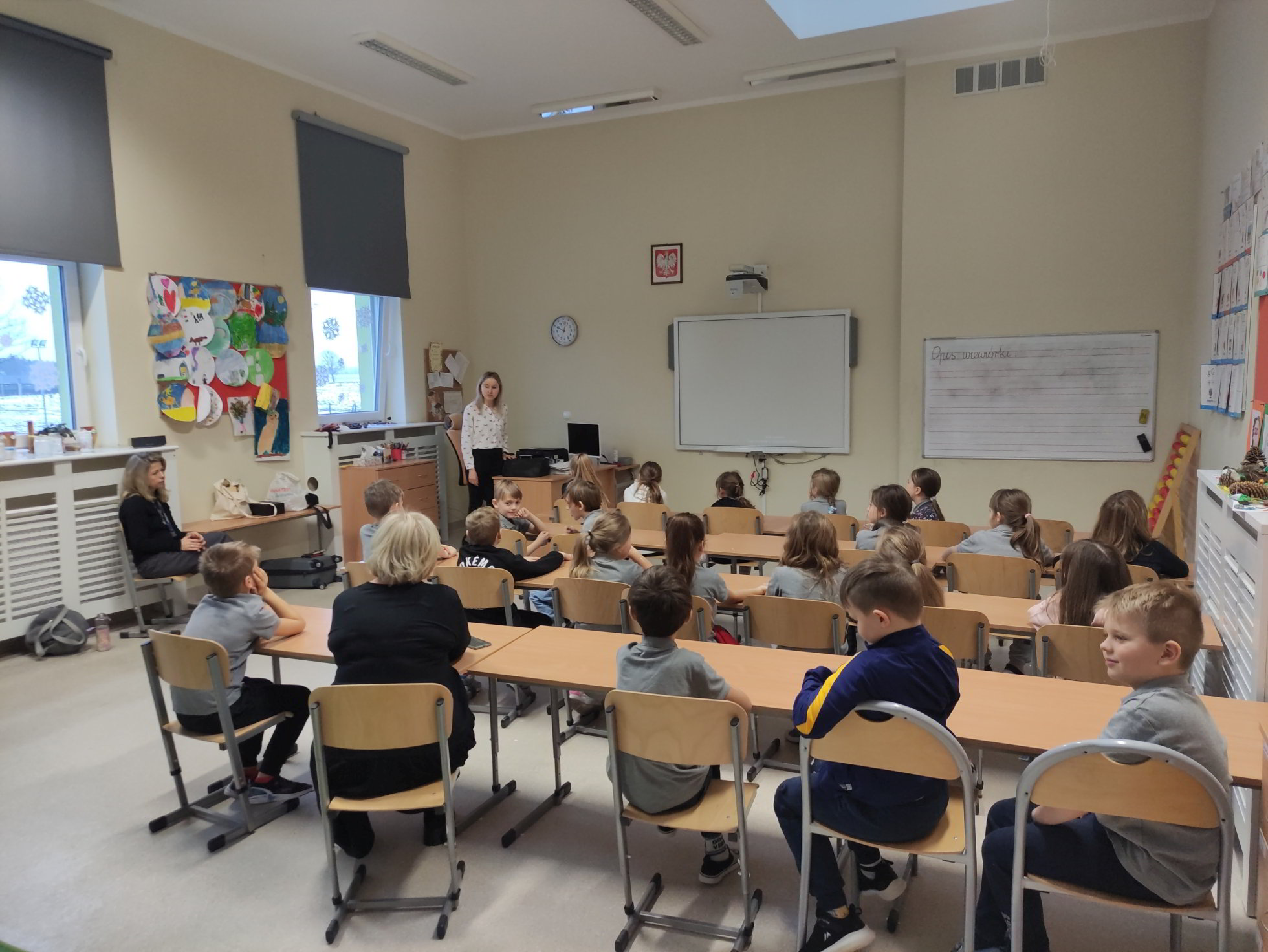 Na zdjęciu widać klasę szkolną podczas zajęć. Uczniowie w wieku szkolnym podstawowym siedzą na drewnianych krzesłach skierowanych w stronę białej tablicy, na której nie ma żadnych zapisków. Są rozmieszczeni w uformowanych rzędach, skupieni na osobie stojącej przed klasą. Po prawej stronie klasy znajduje się kącik z kolorowymi pracami plastycznymi uczniów i tablica z zapisem lekcji. Po lewej stronie widać okna z zasłonami oraz zegar na ścianie. Klasa jest dobrze oświetlona, a atmosfera wydaje się być spokojna i zorganizowana.