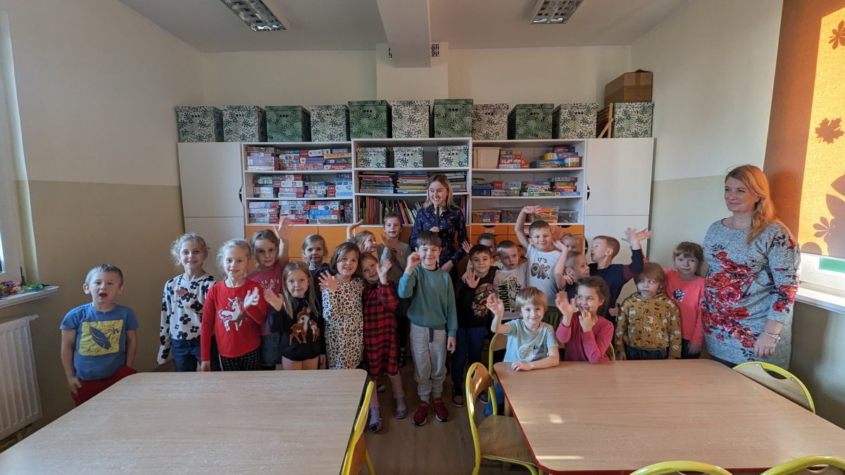 Na zdjęciu widzimy grupę uśmiechniętych dzieci wraz z dwiema dorosłymi kobietami w klasie szkolnej. Dzieci mają różne wyrazy twarzy i pozują do zdjęcia, machając rękami lub pokazując gesty. Są ubrane w codzienne ubrania, a tło za nimi wypełniają półki z książkami i gry edukacyjne. Pomieszczenie jest jasne i kolorowe, z dużym stołem pośrodku, przy którym nie ma osób. Atmosfera jest radosna i beztroska.