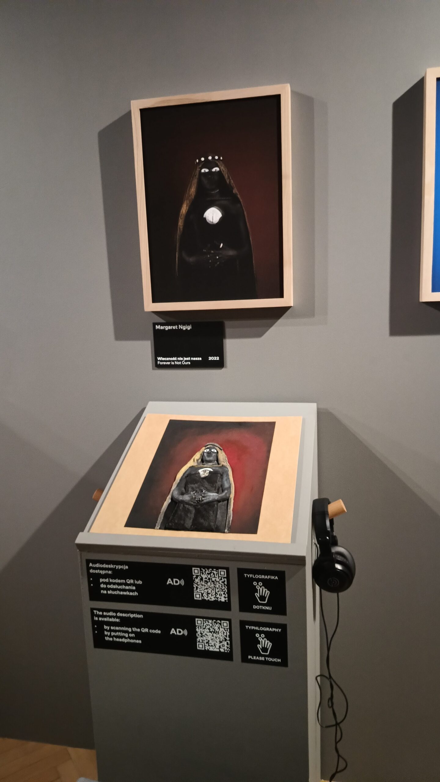 The photo shows Margaret Ngigi's work "Forever is Not Ours" (2022), presented in two versions: the original (on the wall) and a tactile graphic version (below, for touching). The work depicts the figure of a woman with her hand