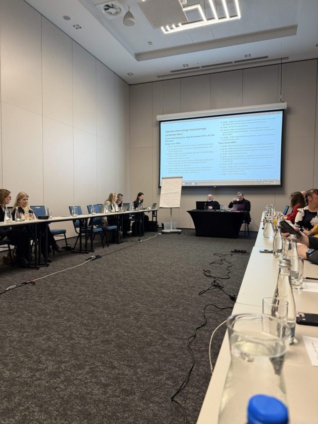 Uczestnicy spotkania zespołu sieci ds. dostępności siedzą przy stołach konferencyjnych i pracują przy laptopach, a na ekranie wyświetlona jest agenda spotkania.