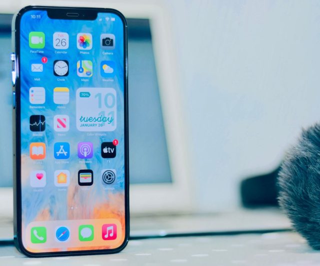 Zdjęcie kolorowe przedstawia współczesny smartfon z dużym, kolorowym ekranem wyświetlającym różnorodne aplikacje. Telefon jest ustawiony pionowo na niezdefiniowanym jasnym tle z widoczną dolną częścią jakiegoś przedmiotu i niewyraźnymi kształtami. Ekran jest wypełniony ikonami aplikacji takich jak FaceTime, kalendarz, zdjęcia, kamera, pogoda, ustawienia i inne. Na ekranie widać również pasek stanu baterii, który pokazuje 70% naładowania. W dolnej części zdjęcia, częściowo niewyraźna, pojawia się sierść zwierzęcia, być może kota, sugerując, że zwierzę to znajduje się tuż obok urządzenia. Dominują kolory są jasne i pastelowe, a całość ma przyjemny, łagodny charakter.