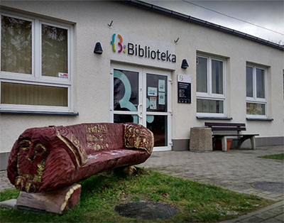 The entrance to a library – a single-storey white building with the word “Biblioteka” (Library) above the door, in front of which stands a carved wooden bench resembling a tree trunk with an eagle motif.