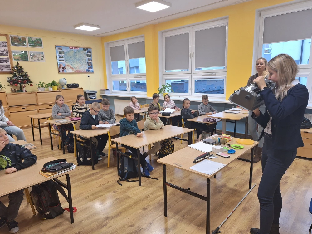Na zdjęciu widać grupę dzieci siedzących w szkolnej klasie, zwróconych w stronę prowadzącej zajęcia kobiety, która trzyma w rękach urządzenie przypominające maszynę brajlowską. W pomieszczeniu znajdują się żółte ściany, okna z roletami oraz elementy dekoracyjne, takie jak choinka i mapa Polski na ścianie. Na ławkach dzieci leżą podręczniki i przybory szkolne, a na stole prowadzącej widoczne są różne przedmioty edukacyjne, m.in. biała laska i drobne gadżety wspierające osoby z niepełnosprawnością wzrokową. Atmosfera wydaje się zaangażowana i edukacyjna.
