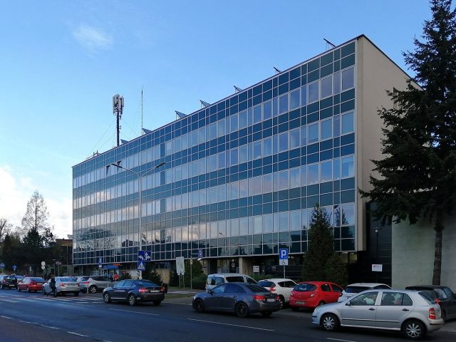 Nowoczesny budynek Okręgowego Szpitala Kolejowego w Katowicach. Sześciokondygnacyjny obiekt z przeszkloną fasadą, z licznymi prostokątnymi oknami. Przed budynkiem zaparkowane są samochody, a na dachu widoczna jest konstrukcja antenowa. Po lewej stronie widać przejście dla pieszych, a po prawej – wysokie drzewa iglaste. Niebo jest bezchmurne.