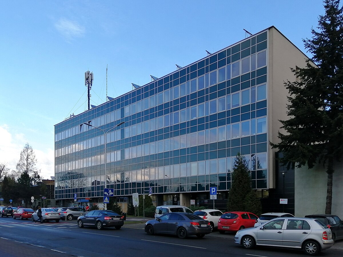 Nowoczesny budynek Okręgowego Szpitala Kolejowego w Katowicach. Sześciokondygnacyjny obiekt z przeszkloną fasadą, z licznymi prostokątnymi oknami. Przed budynkiem zaparkowane są samochody, a na dachu widoczna jest konstrukcja antenowa. Po lewej stronie widać przejście dla pieszych, a po prawej – wysokie drzewa iglaste. Niebo jest bezchmurne.
