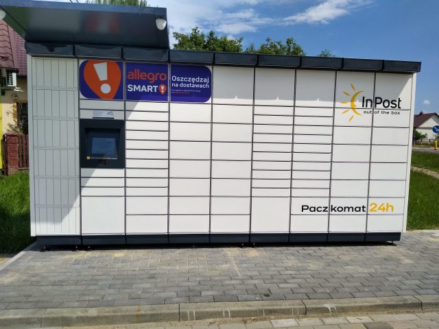 Na zdjęciu widoczny jest automat do odbioru paczek (paczkomat) firmy InPost. Urządzenie to jest umieszczone na zewnątrz i składa się z szeregu małych, zamkniętych skrytek, oznaczonych numerami dla poszczególnych przesyłek. Po lewej stronie znajduje się ekran dotykowy do obsługi systemu oraz instrukcje użytkowania. Automat jest biały z charakterystycznymi pomarańczowo-szarymi akcentami firm InPost i Allegro, a na środku i po prawej stronie ma duże logo InPost. Nad ekranem dotykowym znajduje się logo Allegro Smart, a pod nim hasło promocyjne "Oszczędzaj na dostawach". Z prawej strony, w dolnej części urządzenia umieszczony jest napis "Paczkomat 24h", co wskazuje na całodobową dostępność usługi. Automat stoi na brukowanej nawierzchni, a w tle widać zieloną trawę i niewielki budynek mieszkalny przy słonecznej pogodzie.