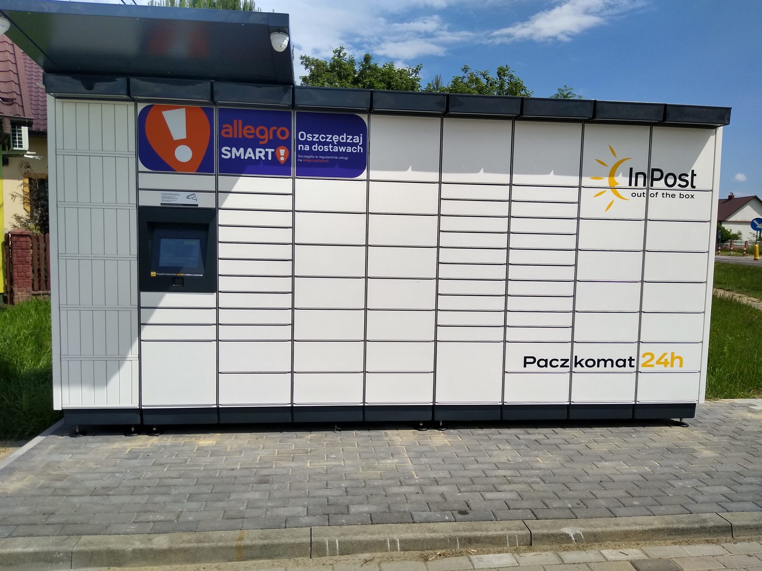 Na zdjęciu widoczny jest automat do odbioru paczek (paczkomat) firmy InPost. Urządzenie to jest umieszczone na zewnątrz i składa się z szeregu małych, zamkniętych skrytek, oznaczonych numerami dla poszczególnych przesyłek. Po lewej stronie znajduje się ekran dotykowy do obsługi systemu oraz instrukcje użytkowania. Automat jest biały z charakterystycznymi pomarańczowo-szarymi akcentami firm InPost i Allegro, a na środku i po prawej stronie ma duże logo InPost. Nad ekranem dotykowym znajduje się logo Allegro Smart, a pod nim hasło promocyjne "Oszczędzaj na dostawach". Z prawej strony, w dolnej części urządzenia umieszczony jest napis "Paczkomat 24h", co wskazuje na całodobową dostępność usługi. Automat stoi na brukowanej nawierzchni, a w tle widać zieloną trawę i niewielki budynek mieszkalny przy słonecznej pogodzie.