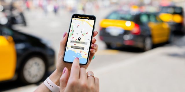 Zdjęcie przedstawia zbliżenie na dłoń osoby trzymającej smartfon. Na ekranie telefonu wyświetlona jest mapa aplikacji do współdzielenia przejazdów, wskazująca na dostępność różnych samochodów w pobliżu. Na ekranie mapy zaznaczone jest wybrane miejsce odbioru oraz opcja zamówienia przejazdu. Tło jest rozmyte, ale widać żółte taksówki, co wskazuje, że scena prawdopodobnie rozgrywa się w miejskim zatłoczonym środowisku. Osoba trzymająca telefon ma na sobie białą opaskę na nadgarstku i pomalowane paznokcie, co sugeruje swobodne i nowoczesne otoczenie.