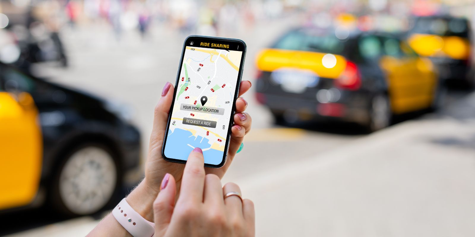 Zdjęcie przedstawia zbliżenie na dłoń osoby trzymającej smartfon. Na ekranie telefonu wyświetlona jest mapa aplikacji do współdzielenia przejazdów, wskazująca na dostępność różnych samochodów w pobliżu. Na ekranie mapy zaznaczone jest wybrane miejsce odbioru oraz opcja zamówienia przejazdu. Tło jest rozmyte, ale widać żółte taksówki, co wskazuje, że scena prawdopodobnie rozgrywa się w miejskim zatłoczonym środowisku. Osoba trzymająca telefon ma na sobie białą opaskę na nadgarstku i pomalowane paznokcie, co sugeruje swobodne i nowoczesne otoczenie.