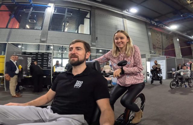 Na zdjęciu widoczny jest mężczyzna w czarnej koszulce z logo "BBS Bike", siedzący w specjalnej rikszy przeznaczonej dla osób z niepełnosprawnością. Kobieta w różowej bluzce w białe kropki stoi za nim, trzymając kierownicę pojazdu i uśmiechając się. W tle znajduje się hala targowa z innymi uczestnikami wydarzenia, stoiskami wystawienniczymi oraz osobami poruszającymi się na wózkach inwalidzkich i innowacyjnych pojazdach wspomagających mobilność.