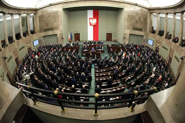 Sala obrad Sejmu RP podczas sesji plenarnej. Posłowie stoją i skupiają uwagę na mównicy, gdzie znajduje się prowadzący obrady. Na ścianie za mównicą wisi duża flaga Polski między dwoma flagami Unii Europejskiej. Pomieszczenie ma półkoliste ustawienie ław, a całość sceny wydaje się uroczysta i oficjalna.